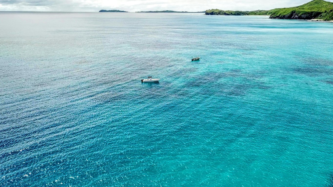 Águas azul-celeste com pequenas embarcações à deriva, cercadas por ilhotas verdes, típicas da tranquilidade e da beleza das Ilhas Fiji.