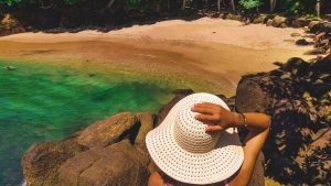 Praia do Português em Ubatuba, uma praia pequena e isolada com águas verdes e rochas ao redor. Uma mulher com chapéu branco aprecia a paisagem.