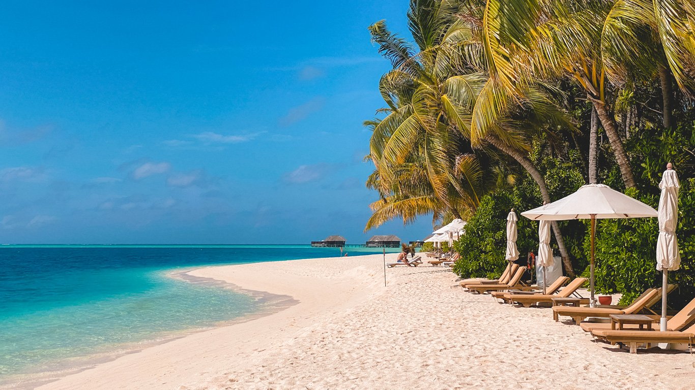 Uma praia paradisíaca nas Maldivas, um dos destinos tropicais mais luxuosos do mundo, com areia branca, coqueiros inclinados e espreguiçadeiras sob guarda-sóis brancos, frente a um mar azul-turquesa sereno.