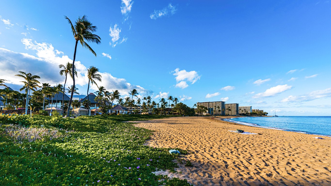 Uma bela praia em Maui, Havaí, com areia dourada se estendendo até o oceano azul sob um céu claro e ensolarado. Palmeiras altas balançam ao vento, cercadas por vegetação verde vibrante e pequenas flores roxas. Ao fundo, resorts luxuosos e edifícios modernos se misturam com a paisagem tropical, enquanto um veleiro navega suavemente pelo mar.