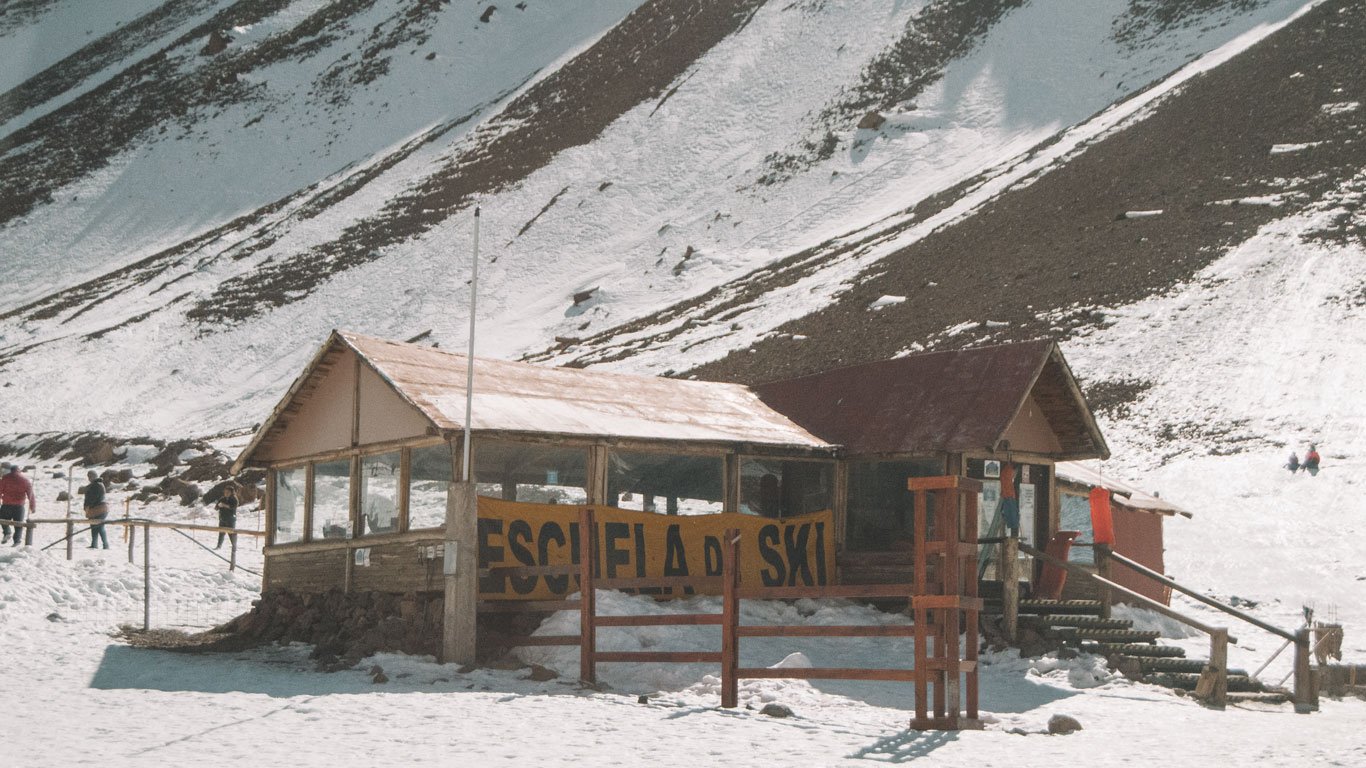 Imagem de uma cabana rústica marcada com a inscrição "Escuela de Ski" em um cenário montanhoso coberto de neve, provavelmente em Mendoza, Argentina. A construção simples, com telhado inclinado e paredes de madeira e vidro, está localizada em meio à neve, cercada por pessoas aproveitando o ambiente. O local parece ser uma escola de esqui, oferecendo um ambiente ideal para atividades de inverno.