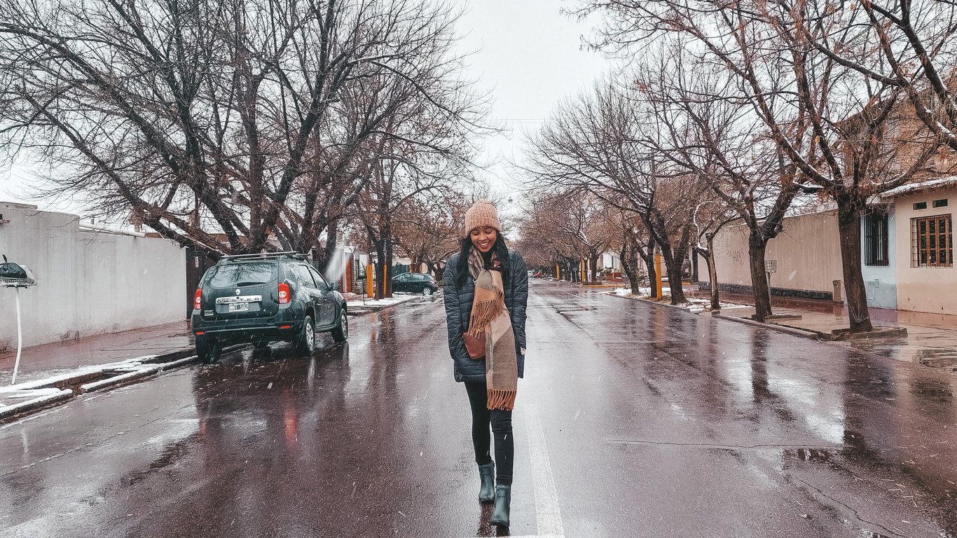 Imagem de uma mulher sorrindo no centro de uma rua residencial em Mendoza, Argentina, durante um dia de neve. Ela está vestida com roupas de inverno, incluindo um gorro de lã, casaco longo e cachecol, enquanto flocos de neve caem suavemente ao redor. A rua molhada é ladeada por árvores com galhos secos e casas ao fundo, criando um ambiente aconchegante e típico de um dia frio na cidade.