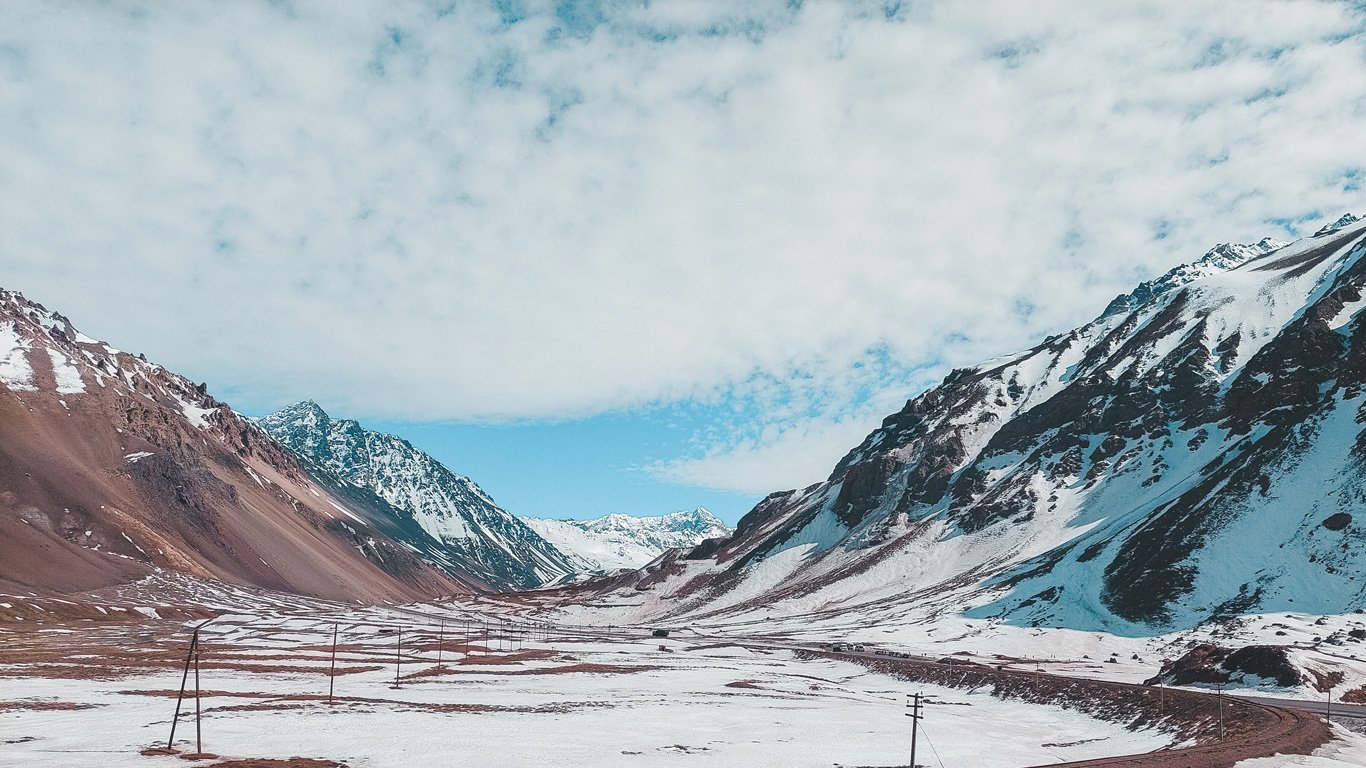 Imagem de um vale cercado por montanhas cobertas parcialmente por neve, provavelmente na região de Mendoza, Argentina. O céu azul com nuvens brancas forma um contraste com as montanhas e o solo nevado, criando um cenário sereno e majestoso. A paisagem transmite a imponência e tranquilidade da natureza andina durante o inverno.