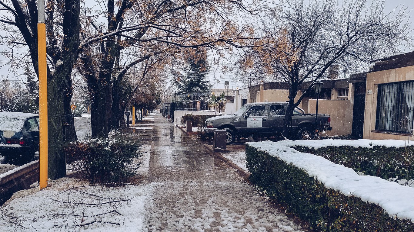 Imagem de uma rua residencial em Mendoza, Argentina, durante um dia de neve. Árvores com galhos secos e parcialmente cobertos por neve ladeiam a calçada molhada, enquanto carros estacionados e uma caminhonete também exibem uma fina camada de neve. O cenário transmite tranquilidade e reflete um típico dia de inverno na cidade.