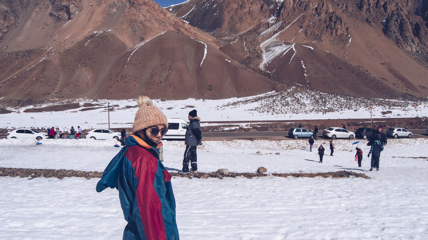 Imagem de uma pessoa vestindo roupas de frio, incluindo um gorro de lã e óculos de sol, em um cenário montanhoso coberto de neve em Mendoza, Argentina. Ao fundo, há outras pessoas aproveitando o ambiente nevado, carros estacionados e uma paisagem com montanhas de tons terrosos e traços de neve. A cena captura um momento descontraído em meio à beleza das montanhas andinas no inverno.