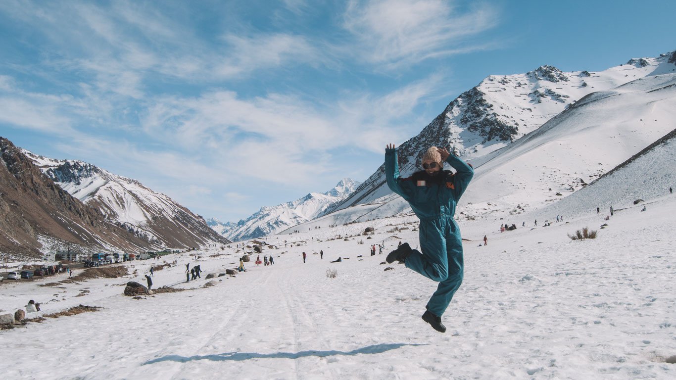 Imagem de uma pessoa saltando em um cenário montanhoso coberto de neve, provavelmente na região de Mendoza, Argentina. Ao fundo, há montanhas majestosas, várias pessoas aproveitando a neve, e um céu azul claro com poucas nuvens. A cena transmite alegria e um ambiente perfeito para atividades de inverno.