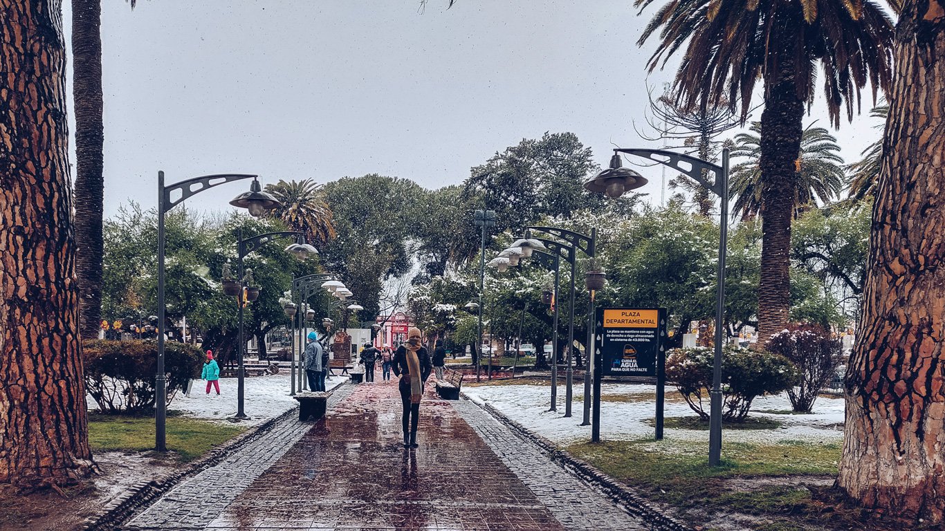 Imagem de uma praça arborizada durante um dia de neve em Mendoza, Argentina. No centro, há uma pessoa caminhando em um caminho de tijolos molhados, cercado por postes de iluminação e árvores altas, incluindo palmeiras. Ao fundo, é possível ver outras pessoas apreciando o ambiente nevado, com vegetação parcialmente coberta pela neve, criando uma atmosfera encantadora de inverno urbano.