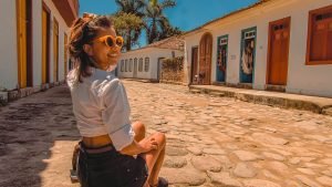 Mulher sorridente usando óculos de sol laranja sentada em uma rua de pedra do centro histórico de Paraty, com casas coloniais coloridas ao fundo.