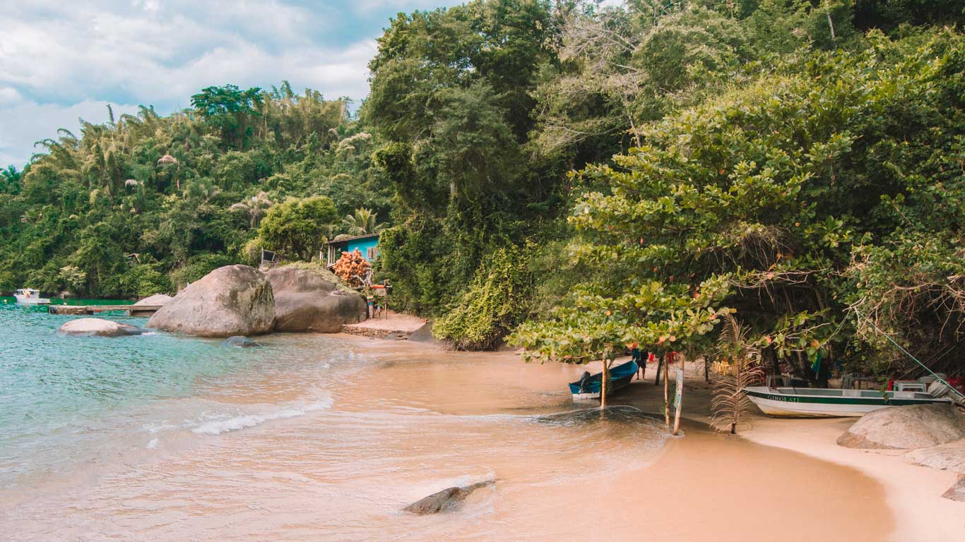 Praia Saco da Velha em Paraty, com faixa de areia ampla e dois visitantes caminhando próximo ao mar, cercada por montanhas verdes.