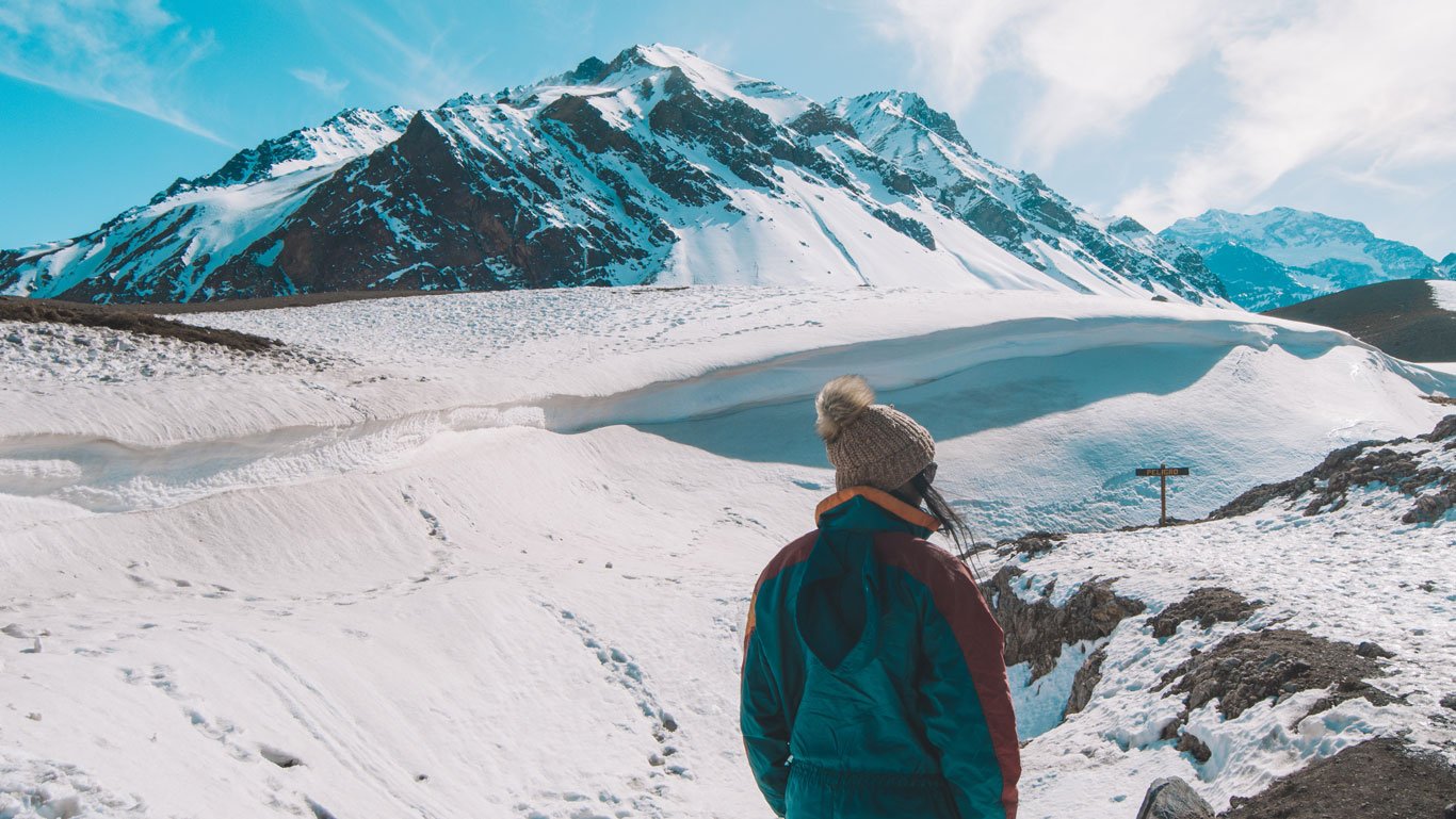 Imagem de uma pessoa de costas, vestindo um casaco de inverno e um gorro de lã, admirando um cenário montanhoso coberto de neve no Parque Aconcágua, em Mendoza, Argentina. Ao fundo, destacam-se as imponentes montanhas dos Andes com picos nevados e um céu azul claro, enquanto o terreno apresenta marcas de pegadas na neve. A cena transmite uma sensação de tranquilidade e conexão com a natureza em um ambiente gelado e majestoso.