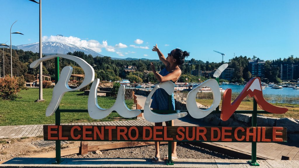 Mulher posando atrás de um letreiro escrito Pucón, apontando para o vulcão Villarrica ao fundo.