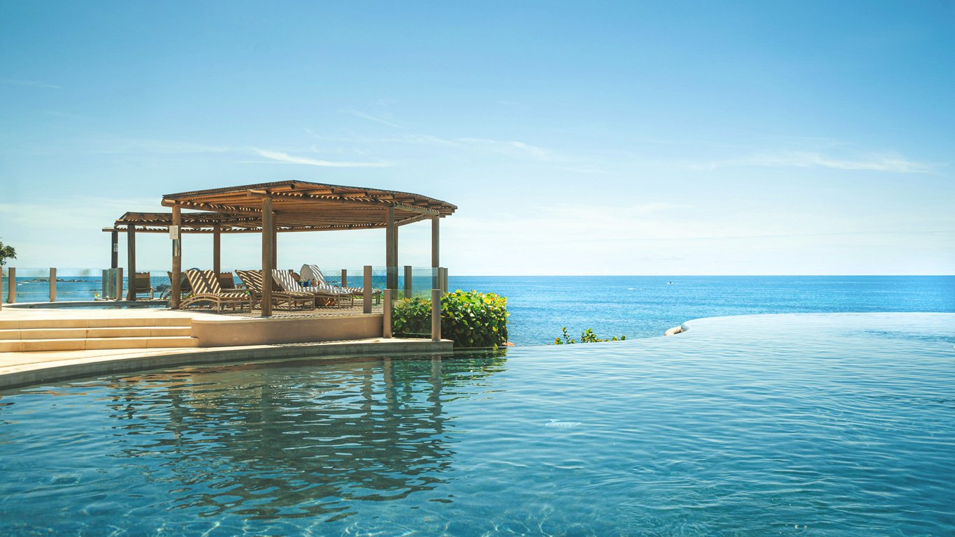 Uma luxuosa piscina de borda infinita em Punta Mita, com vista direta para o oceano azul sob um céu claro. Ao lado da piscina, há um elegante pergolado de madeira com espreguiçadeiras dispostas para relaxamento. O cenário transmite uma sensação de tranquilidade e sofisticação, com a água da piscina se misturando visualmente com o mar ao fundo.