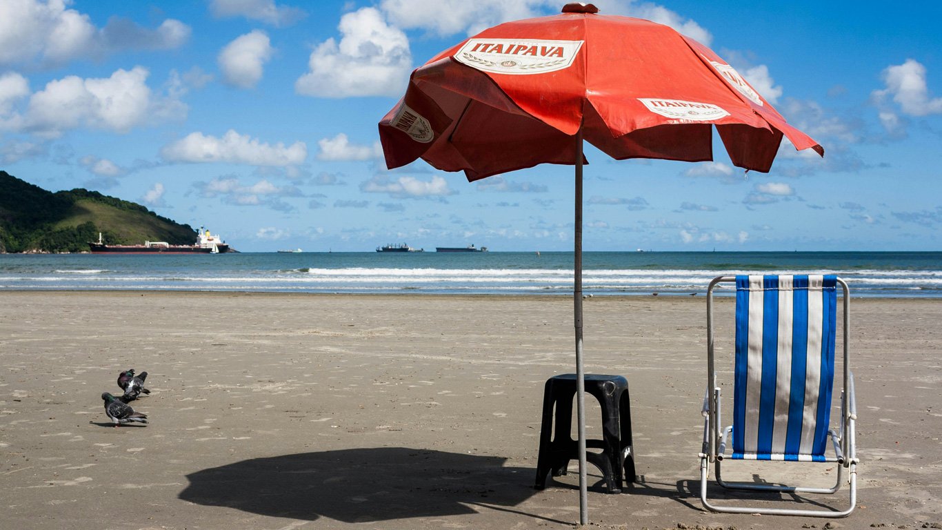 Praia urbana de Santos, com guarda-sol vermelho estampado com o logo de uma marca e uma cadeira de praia azul. O mar ao fundo tem navios ancorados, e a paisagem traz um ar de lazer e descanso.