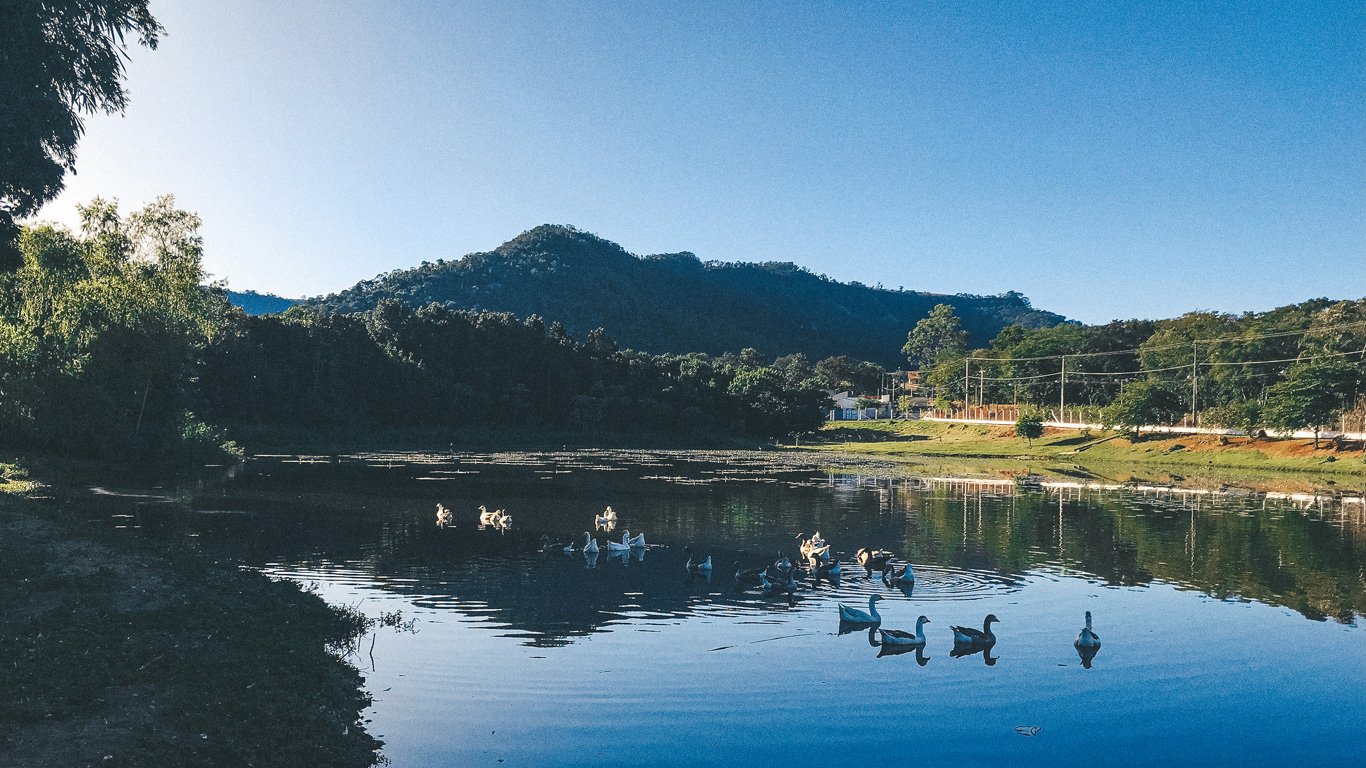 Lago sereno em São Pedro, com patos flutuando na água, cercado por montanhas e vegetação verde. Um lugar ideal para relaxar em meio à natureza.