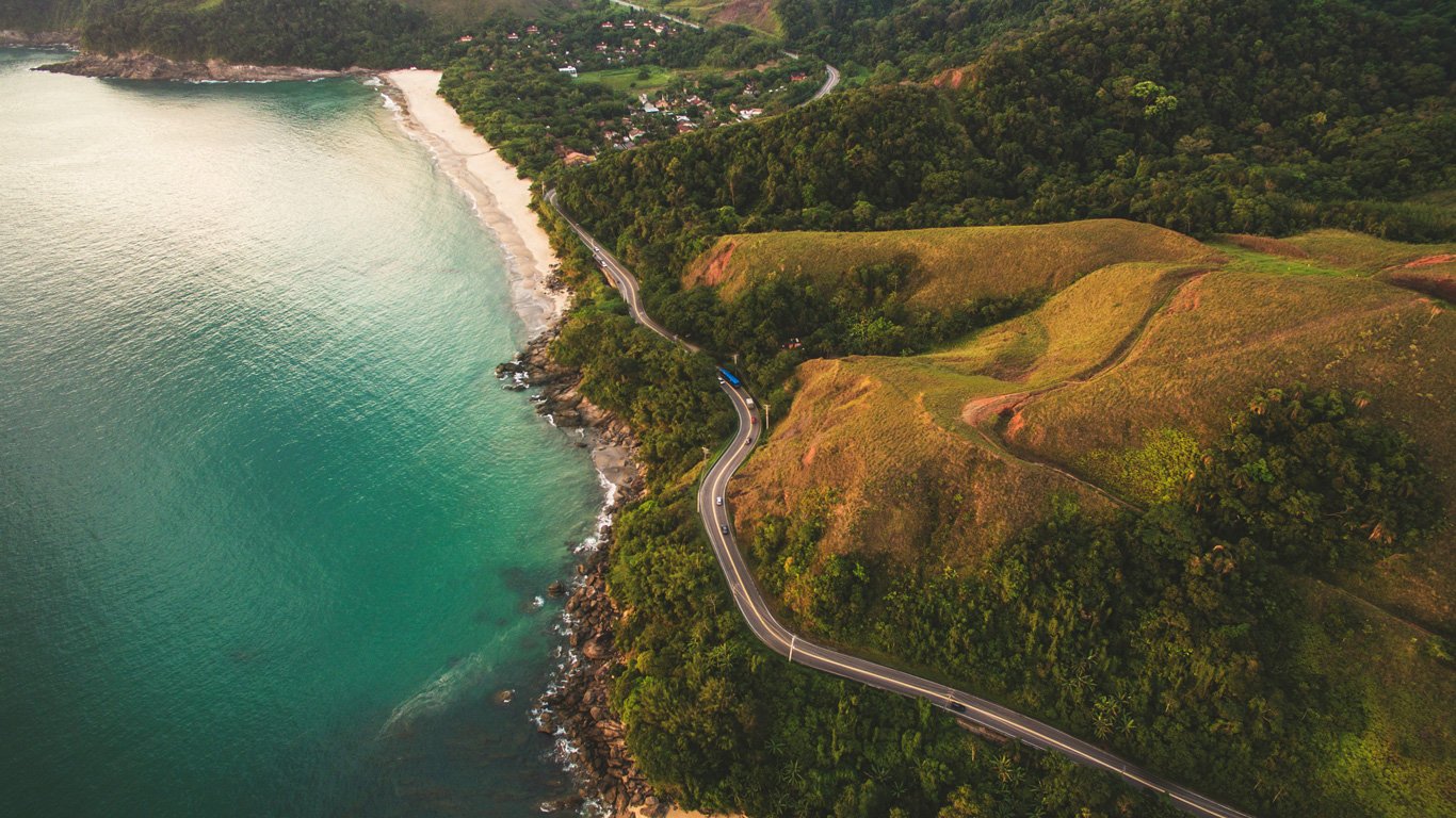 Vista aérea de Toque Toque Pequeno, com uma impressionante estrada sinuosa margeando o oceano azul-esverdeado. O contraste entre o mar, a praia e as colinas cria um visual deslumbrante.