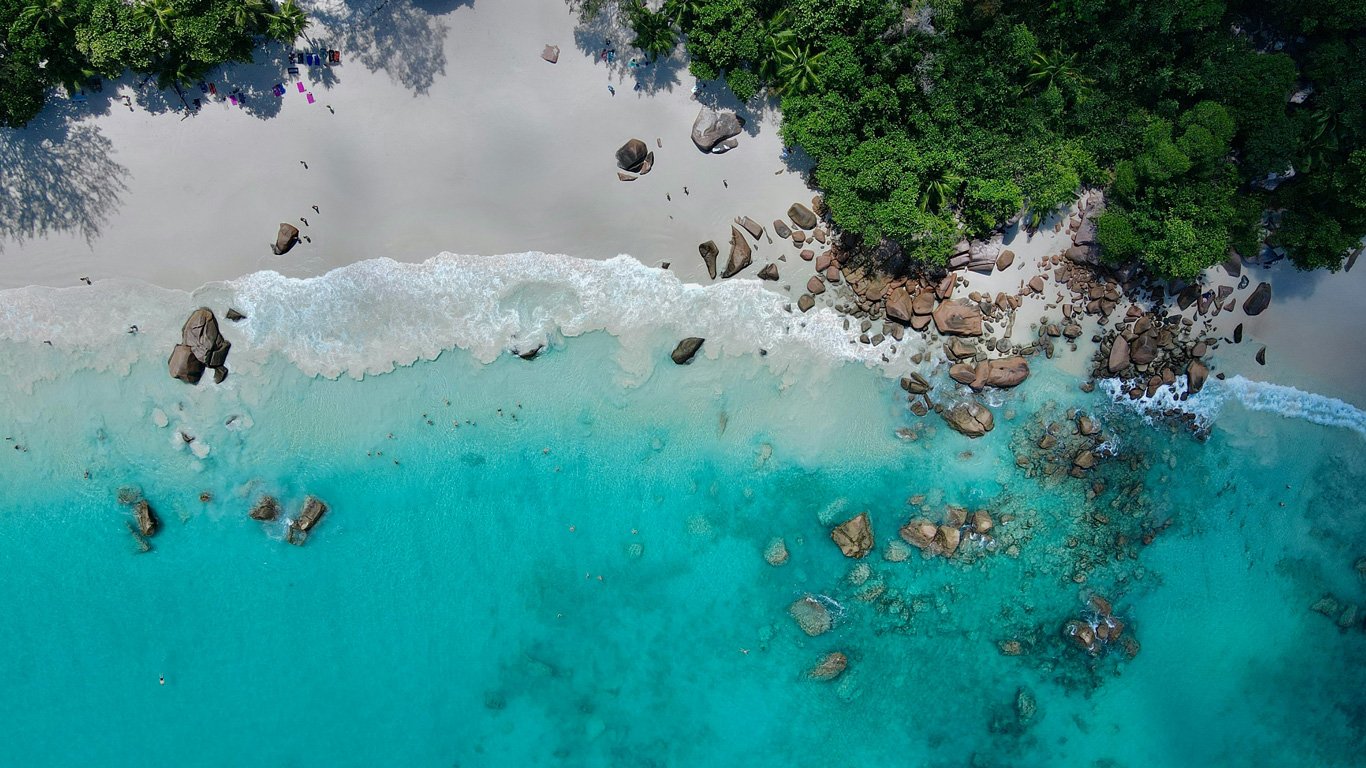 Uma vista aérea deslumbrante de uma praia tropical, onde as águas azul-turquesa encontram a areia branca e imaculada. A praia é cercada por uma vegetação verde exuberante, enquanto grandes rochas espalhadas formam um litoral natural e rústico. Ondas suaves quebram na areia, e algumas pessoas aproveitam o local, relaxando na praia ou nadando na água cristalina.