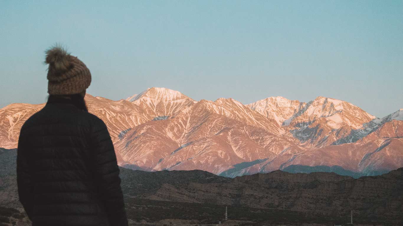 Uma pessoa vestindo roupas de frio observa a grandiosidade das montanhas dos Andes, parcialmente cobertas de neve, sob a luz dourada do pôr do sol. A paisagem montanhosa de Mendoza, Argentina, revela tons terrosos e brancos, criando um contraste impressionante com o céu claro. A cena transmite a imensidão e a beleza da natureza na região de alta montanha.