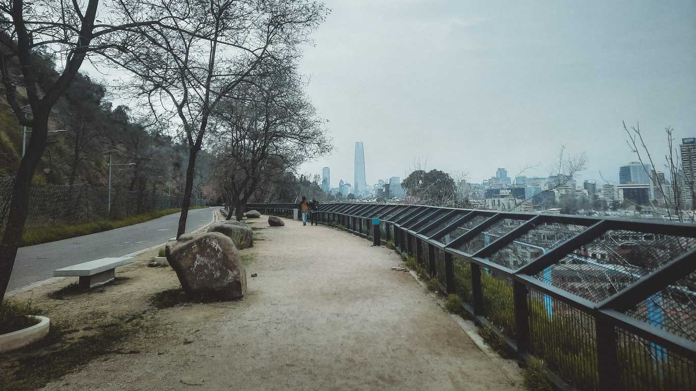 Entrada do Cerro San Cristóbal no bairro Bellavista, em Santiago, com uma trilha arborizada e a cidade ao fundo.