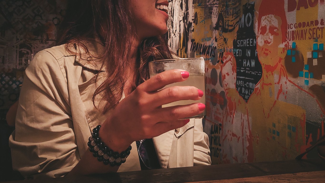 Uma mulher segurando um coquetel em um bar com uma parede cheia de arte urbana ao fundo. O ambiente tem iluminação quente e um clima descontraído.