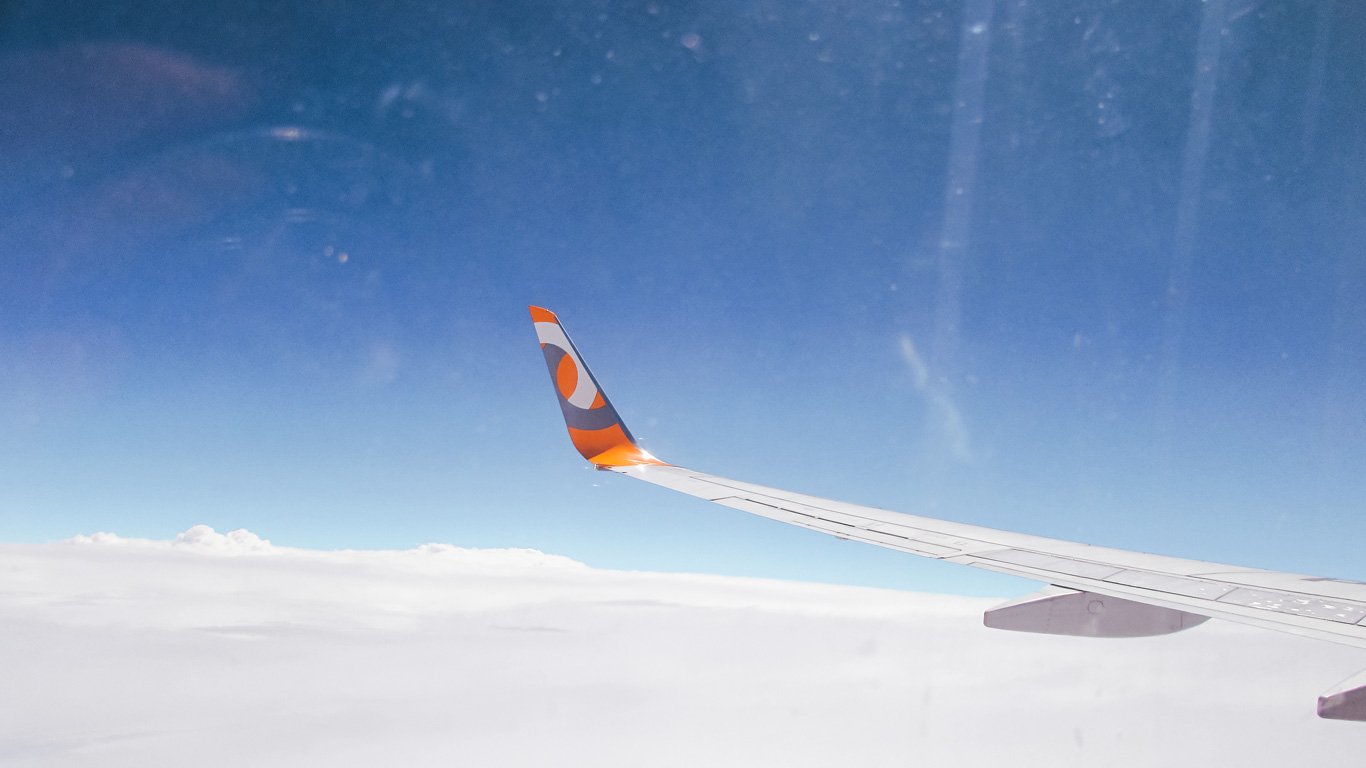 Fotografia da asa de um avião com uma logo laranja e branca na ponta, voando acima de uma camada de nuvens brancas.