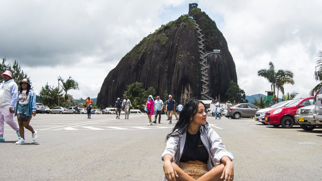 Mulher sentada no estacionamento da pedra El Peñol posando para a foto, ao fundo a grandiosa pedra El Peñol.