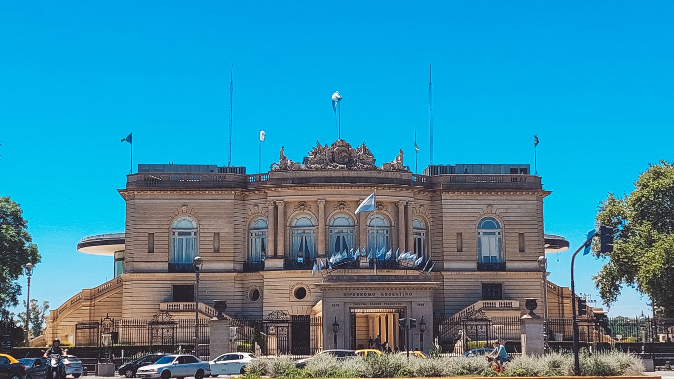 Fachada imponente do Hipódromo Argentino de Palermo, em Buenos Aires, com sua arquitetura clássica e bandeiras da Argentina tremulando sob um céu azul vibrante.