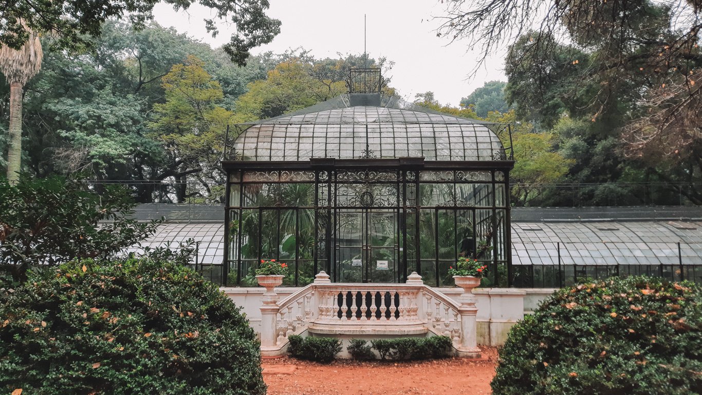 Uma bela estufa de vidro e ferro cercada por vegetação exuberante no Jardim Botânico de Buenos Aires, transmitindo um ambiente sereno e natural.