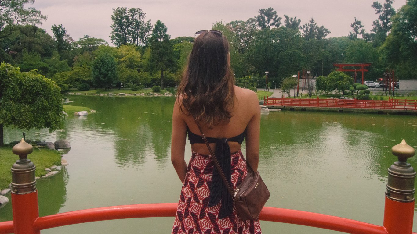 Uma mulher de costas, vestindo uma saia estampada e um top preto, aprecia a paisagem tranquila do Jardim Japonês de Buenos Aires. O lago verde-reflexivo e as estruturas vermelhas dão um toque oriental ao cenário.