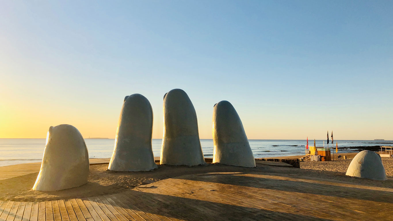 Escultura "La Mano" na Praia Brava, em Punta del Este, Uruguai, capturada ao nascer do sol. As cinco enormes pontas de dedos emergem da areia dourada, simbolizando a presença humana e se destacando contra o céu azul claro e o oceano tranquilo ao fundo. A luz suave do amanhecer ilumina a cena, criando um contraste entre as sombras da escultura e o brilho dourado da praia.