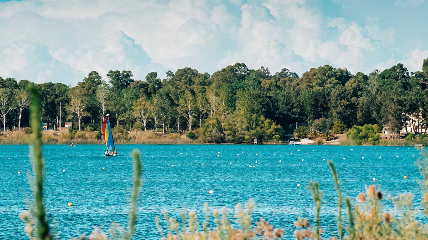 Paisagem serena da Laguna del Sauce, em Punta del Este, Uruguai, com águas azul-turquesa refletindo o céu. Ao fundo, uma densa vegetação de árvores verdes contorna a margem, criando um ambiente natural e tranquilo. Um pequeno veleiro com vela colorida navega suavemente pelo lago, adicionando um toque vibrante à cena.