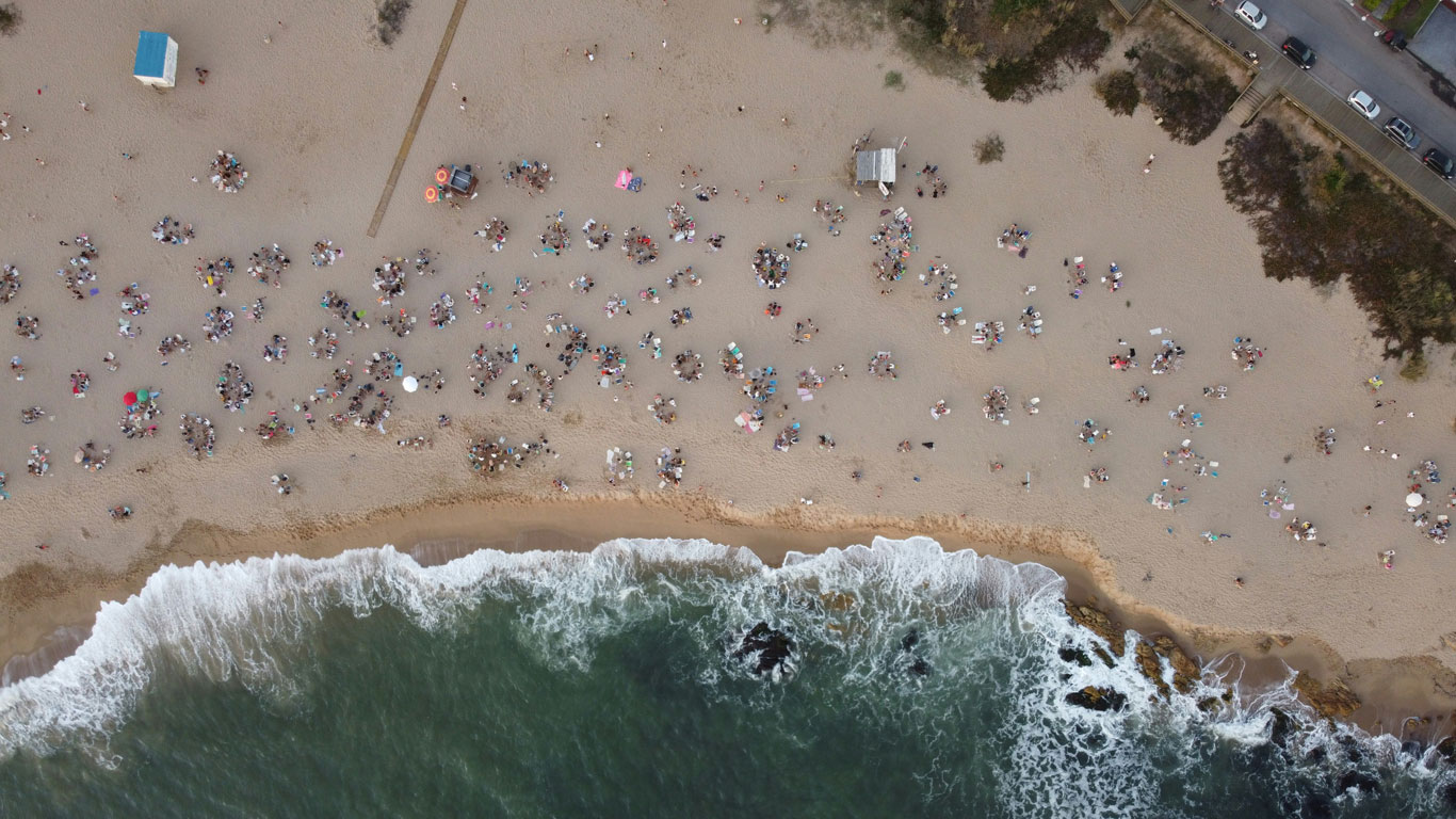 Vista aérea da praia de Manantiales, em Punta del Este, Uruguai, mostrando a areia dourada repleta de banhistas organizados em pequenos grupos. O mar de águas esverdeadas e espumantes encontra a costa, formando ondas suaves que se quebram na praia. A imagem captura a vibração do verão, com guarda-sóis coloridos, esteiras e uma atmosfera animada à beira-mar.