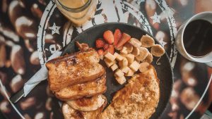 Uma refeição de café da manhã servida em um prato preto sobre uma mesa com estampa de grãos de café. O prato contém fatias douradas de rabanada com calda, uma omelete temperada e frutas frescas, incluindo morangos, banana fatiada e pedaços de abacaxi. Ao lado, há um copo de café gelado e uma xícara de café preto quente.