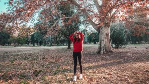 Uma mulher vestindo um moletom vermelho e calça preta posa sob uma grande árvore com folhas alaranjadas, típicas do outono. O chão está coberto por folhas secas, enquanto o fundo do parque exibe mais árvores e um céu azul suave. A cena transmite uma atmosfera tranquila e aconchegante, capturando a essência do outono em Mendoza, Argentina.