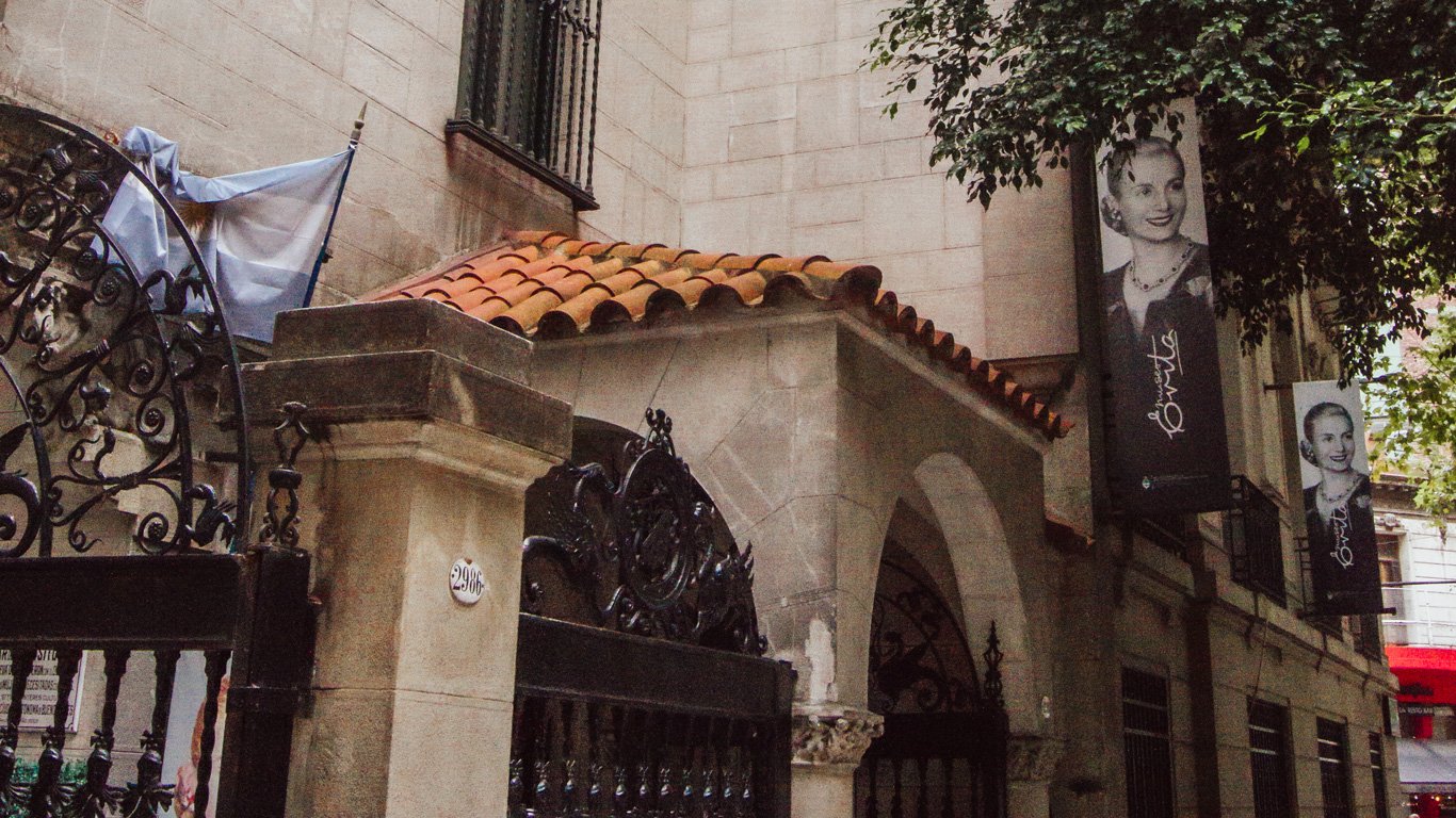 Entrada do Museu Evita em Palermo, Buenos Aires, com sua arquitetura tradicional e portões ornamentados. Banners com a imagem icônica de Eva Perón decoram a fachada.