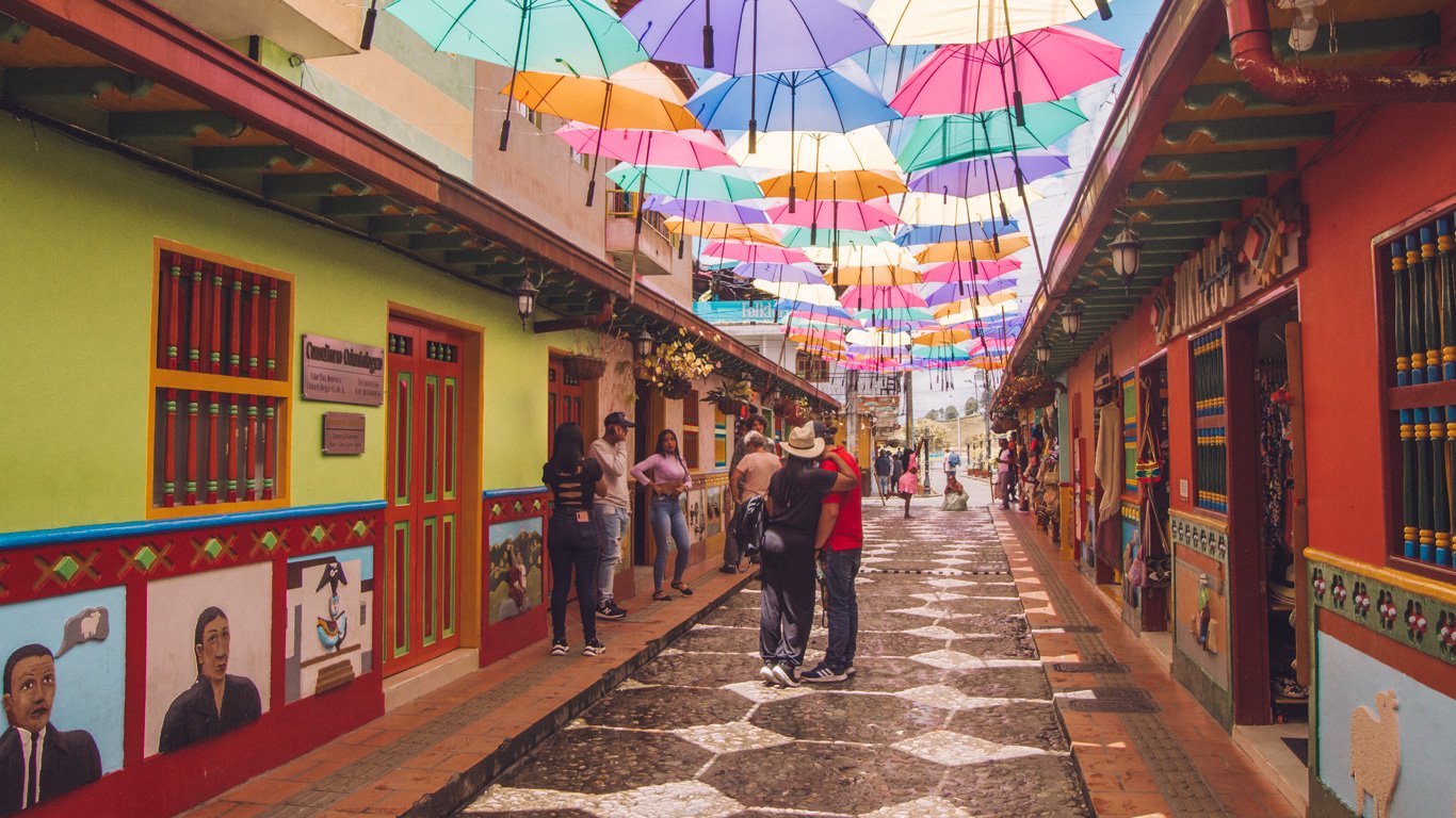 Rua com diversos guada-chuvas coloridos suspensos. As casas são coloridas e diversas pessoas estão andando descontraídas pelo lugar.