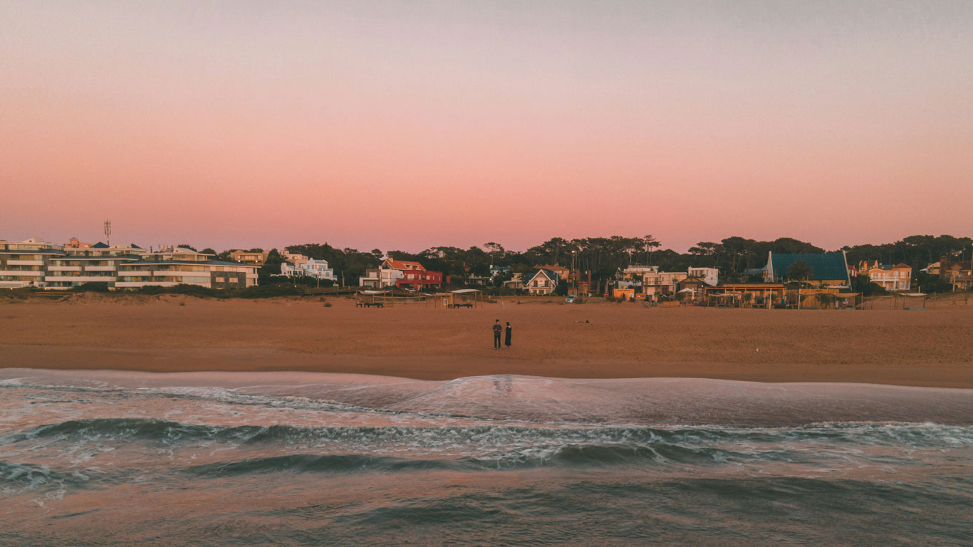 Pôr do sol em La Barra, Punta del Este, Uruguai, com o céu tingido de tons rosados e alaranjados. O mar calmo e as ondas suaves contrastam com a extensa faixa de areia dourada, onde duas pessoas caminham tranquilamente. Ao fundo, casas coloridas e modernos edifícios refletem o charme e a exclusividade da região.