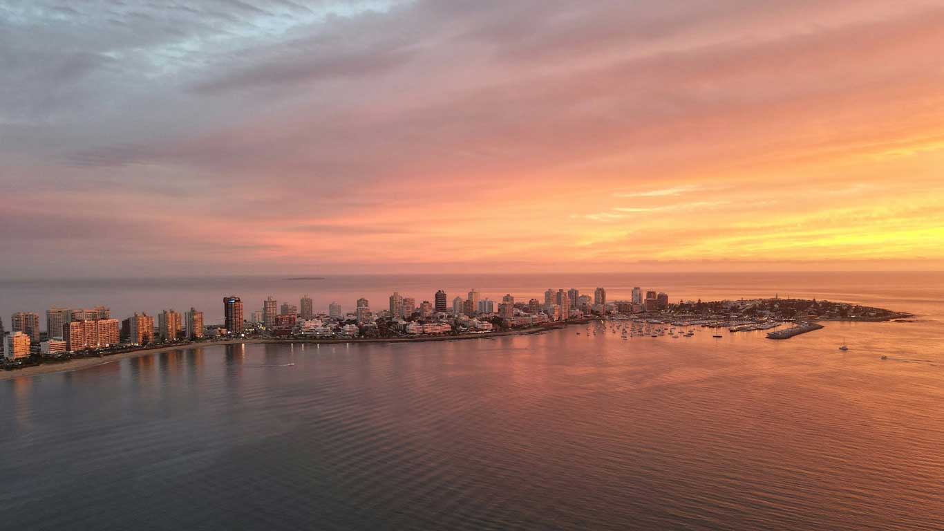 Vista aérea de Punta del Este, Uruguai, durante um pôr do sol espetacular. O céu exibe tons alaranjados e rosados refletidos nas águas calmas, enquanto a cidade com seus edifícios modernos se estende ao longo da costa. O porto e os barcos ancorados completam a paisagem desse famoso destino turístico da América do Sul.