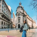 Uma mulher de óculos escuros está posando em uma rua ampla e ensolarada, cercada por edifícios históricos ornamentados, no centro de Santiago do Chile, um ótimo local para considerar quando pensar em onde ficar em Santiago.
