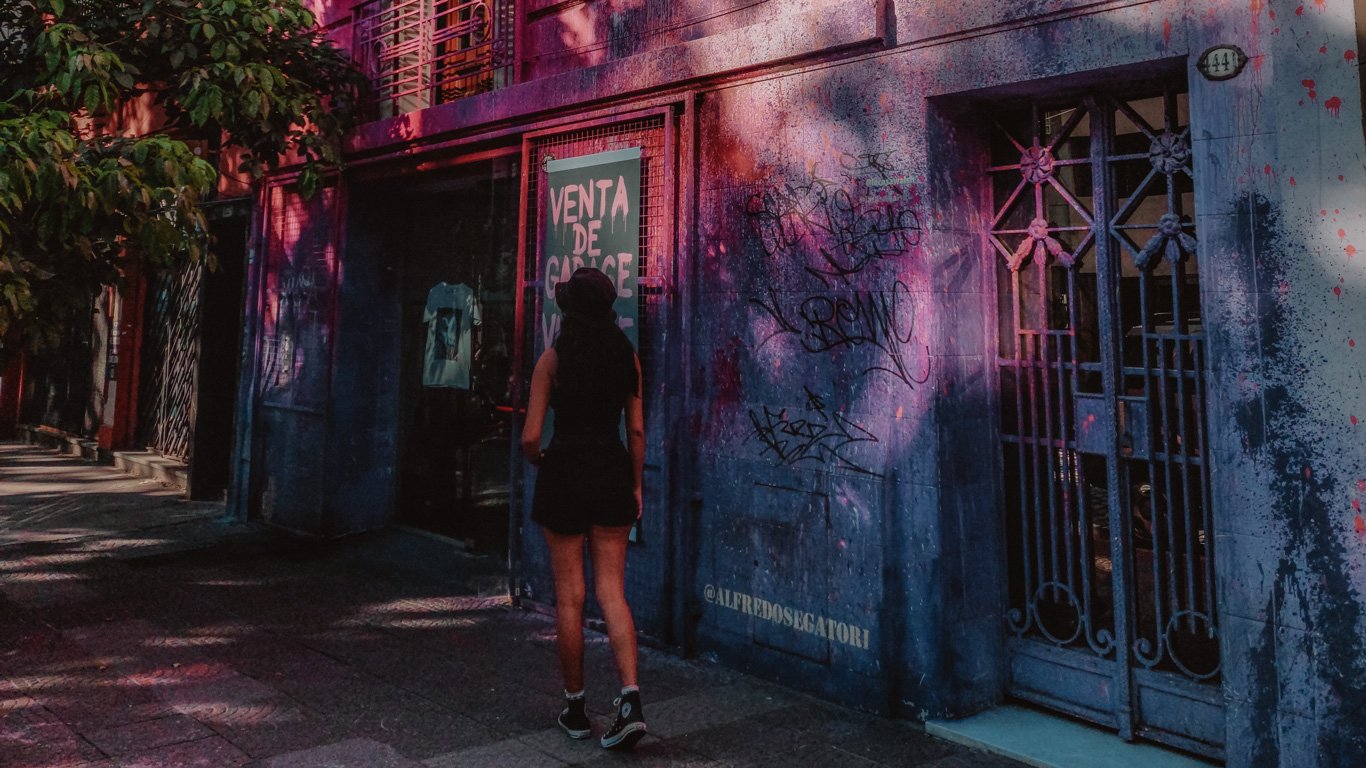 Uma mulher vestida de preto, com um chapéu e tênis, observa a fachada de um prédio pintado em tons escuros com detalhes em grafite e manchas de tinta colorida no bairro de Palermo, Buenos Aires. Um cartaz na vitrine anuncia uma "Venta de Garage" (Venda de Garagem), sugerindo um evento ou mercado local.