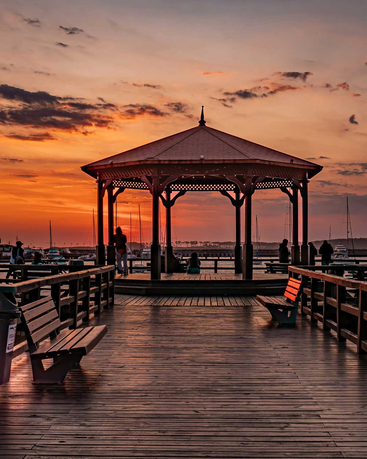 Pôr do sol na Península de Punta del Este, Uruguai, visto de um píer de madeira com um charmoso gazebo central. O céu exibe tons alaranjados e rosados, refletidos na água tranquila do porto, onde barcos estão ancorados. Algumas pessoas aproveitam o momento, sentadas nos bancos ou apreciando a paisagem de pé, criando uma atmosfera serena e romântica.