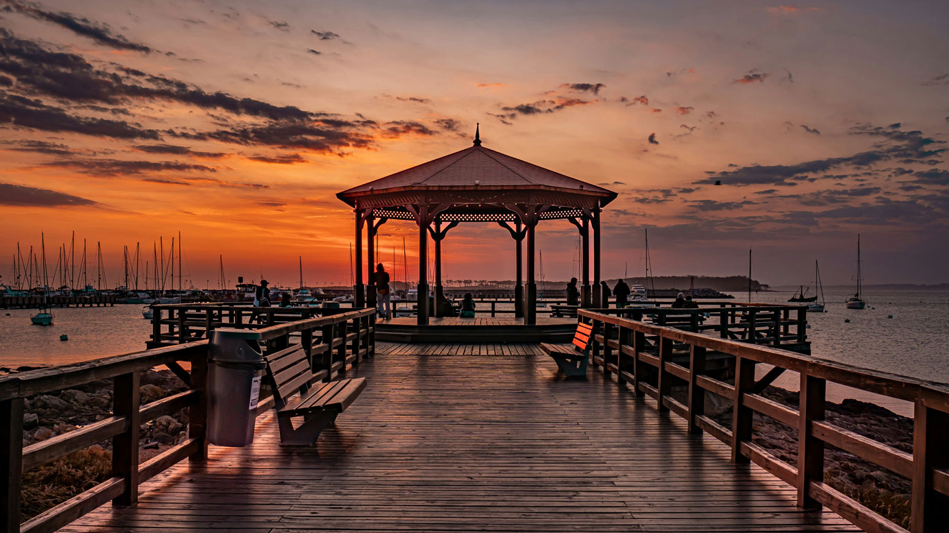 Pôr do sol na Península de Punta del Este, Uruguai, visto de um píer de madeira com um charmoso gazebo central. O céu exibe tons alaranjados e rosados, refletidos na água tranquila do porto, onde barcos estão ancorados. Algumas pessoas aproveitam o momento, sentadas nos bancos ou apreciando a paisagem de pé, criando uma atmosfera serena e romântica.