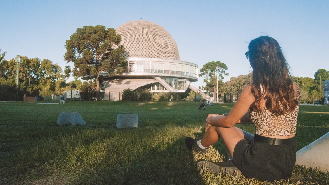 Uma mulher sentada na grama, observando o icônico Planetário Galileo Galilei em Buenos Aires. A estrutura futurista e o céu claro criam um cenário impressionante.