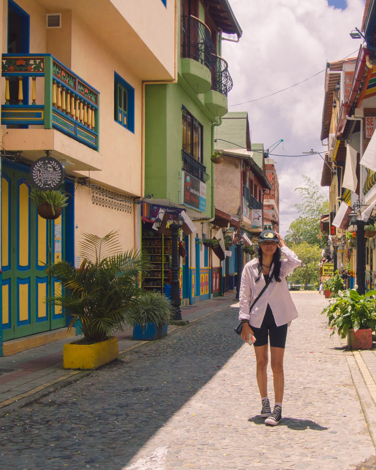 Mulher usando boné, camisa branca e short preto, posa para foto em rua colorida do centro do Pueblo de Guatapé.