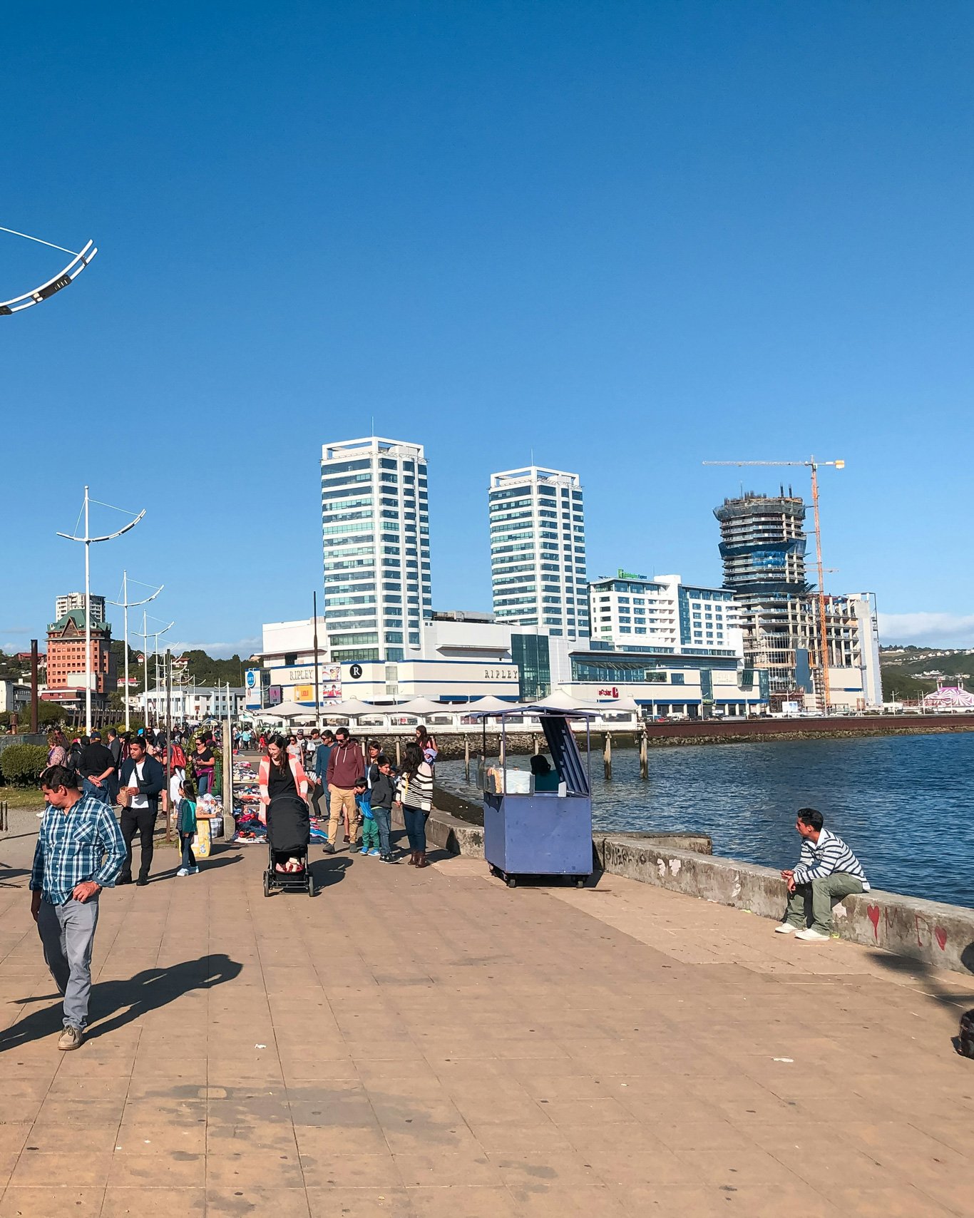 Vista da Costanera de Puerto Montt, com pessoas caminhando e artistas vendendo suas artes. Ao fundo alguns prédios modernos e o shopping Mall Paseo Costanera.