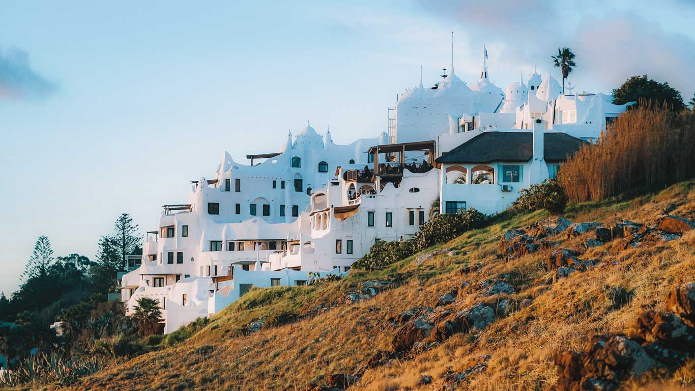 Casapueblo, em Punta Ballena, Punta del Este, Uruguai, com sua arquitetura branca e curvilínea inspirada em Gaudí, iluminada pela luz dourada do entardecer. O edifício icônico, que combina hotel, museu e galeria de arte, destaca-se sobre a encosta rochosa coberta de vegetação seca. O céu azul suave e as sombras alongadas realçam a atmosfera tranquila e artística do local.