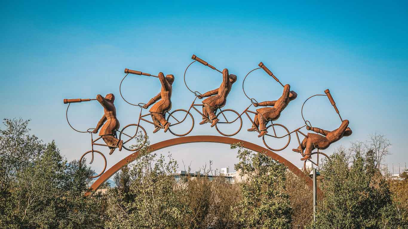 Escultura artística em arco no Parque Bicentenário, Vitacura, Chile, retratando quatro figuras humanas pedalando bicicletas enquanto seguram lunetas. As silhuetas metálicas parecem estar em movimento, criando um efeito dinâmico contra o céu azul. A obra se destaca sobre a vegetação verde ao fundo, adicionando um toque surrealista à paisagem urbana.