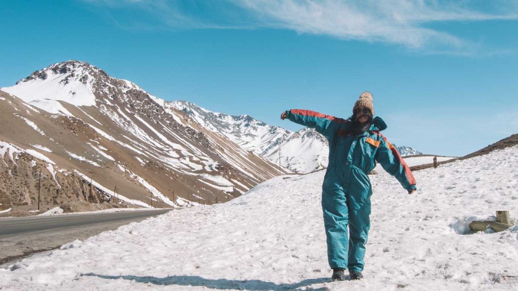 Uma mulher vestindo um macacão de neve azul e vermelho e um gorro de lã posa animada em uma paisagem montanhosa coberta de neve em Mendoza, Argentina. O céu azul claro destaca os picos nevados ao fundo, enquanto a estrada ao lado adiciona profundidade à cena. Ela abre os braços, expressando alegria e energia em meio ao frio da região.