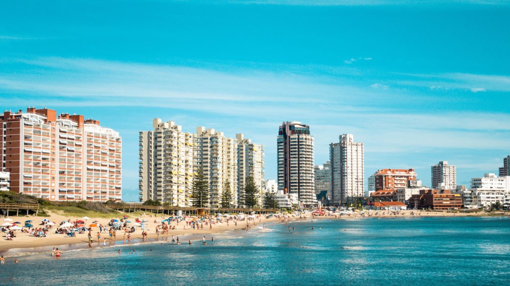 A imagem mostra uma praia movimentada em Punta del Este, Uruguai, com diversas pessoas aproveitando o dia ensolarado à beira-mar. Há guarda-sóis coloridos espalhados pela areia, enquanto o mar azul reflete o céu claro. Ao fundo, arranha-céus modernos e prédios residenciais compõem o horizonte, destacando a fusão entre a natureza e a urbanização deste famoso destino turístico.