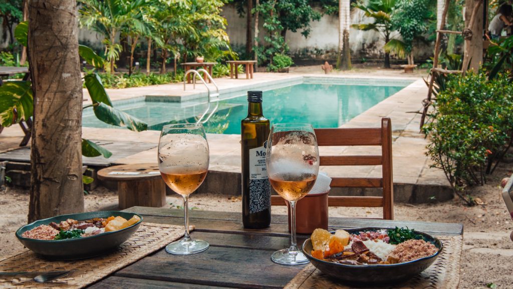 Almoço ao ar livre com duas tigelas de feijoada vegana acompanhadas de arroz, farofa, couve, vinagrete e laranja, servidas com vinho rosé e azeite, em mesa rústica com vista para piscina cercada de verde – sugestão perfeita para quem busca onde comer em Pipa.
