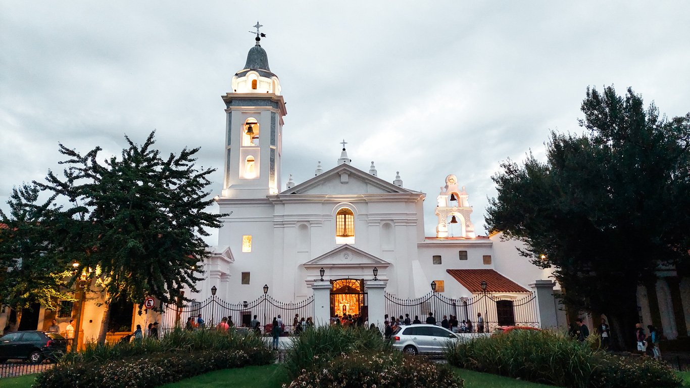 Basílica-Nuestra-Senora-del-Pilar Fachada da Basílica Nuestra Señora del Pilar com torres iluminadas ao entardecer, cercada por jardins e pessoas na entrada. A arquitetura colonial e o céu nublado criam um ambiente acolhedor e histórico.